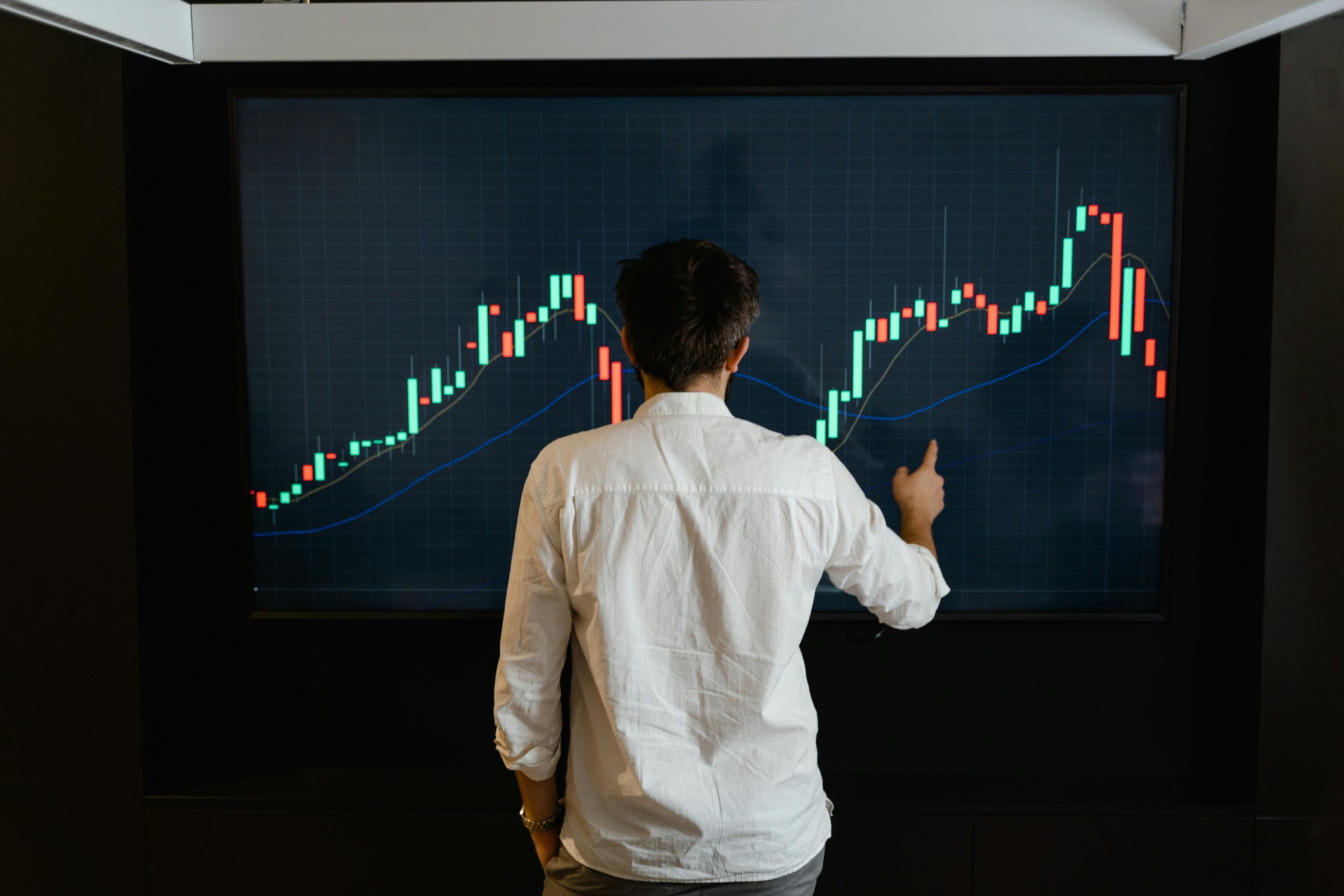 Adult male reviewing stock, FOREX market data on a large display screen indoors.